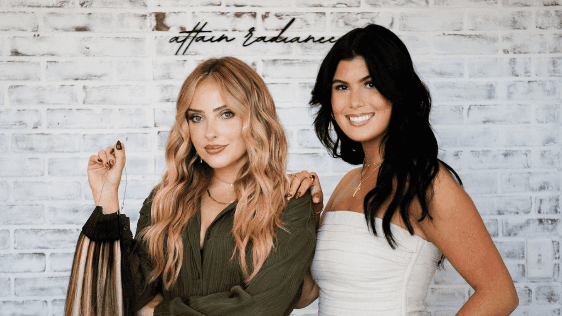 Two women smiling in front of a white brick wall with cursive text above.
