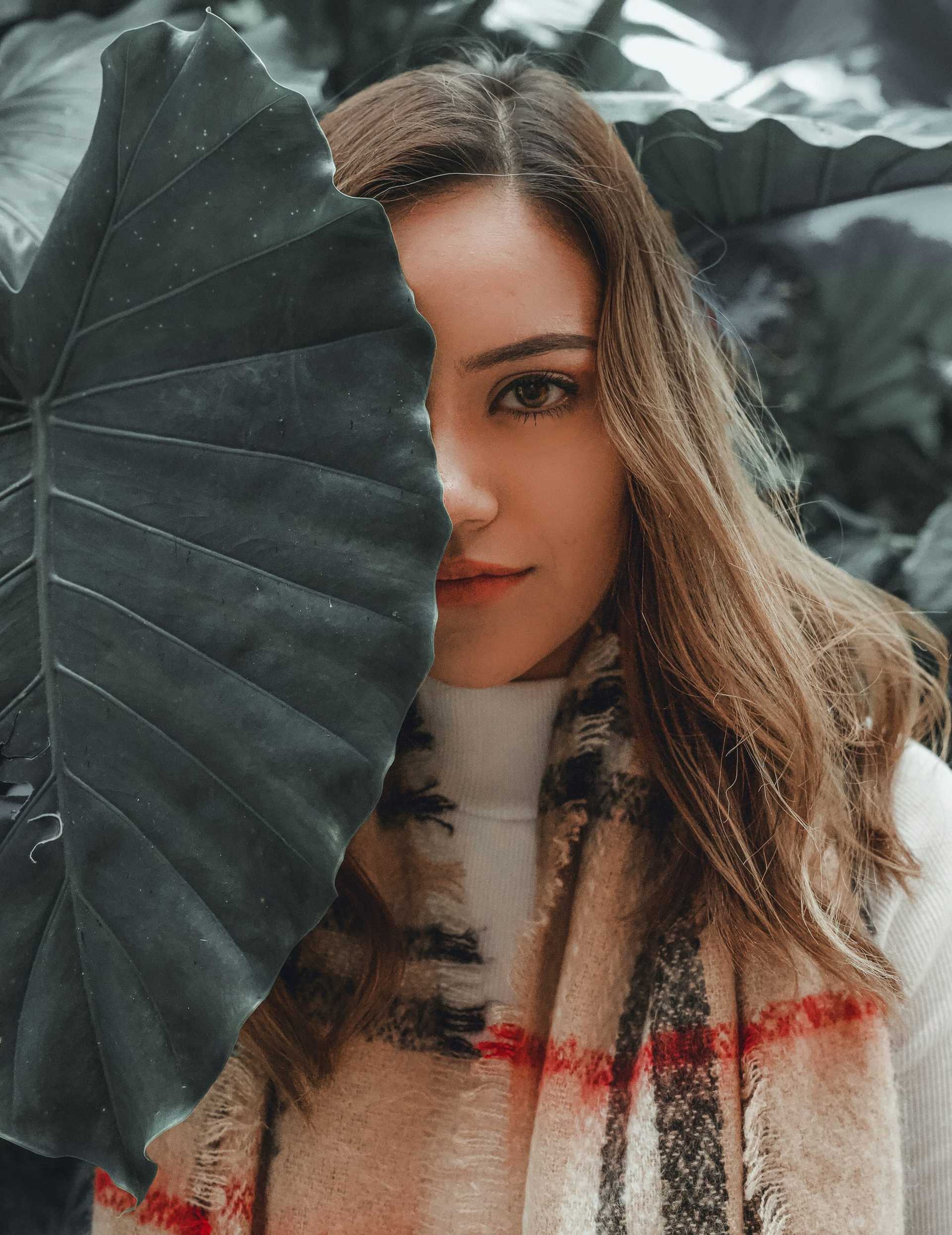 Woman partially hidden by large leaf, wearing a plaid scarf, with lush greenery in the background.