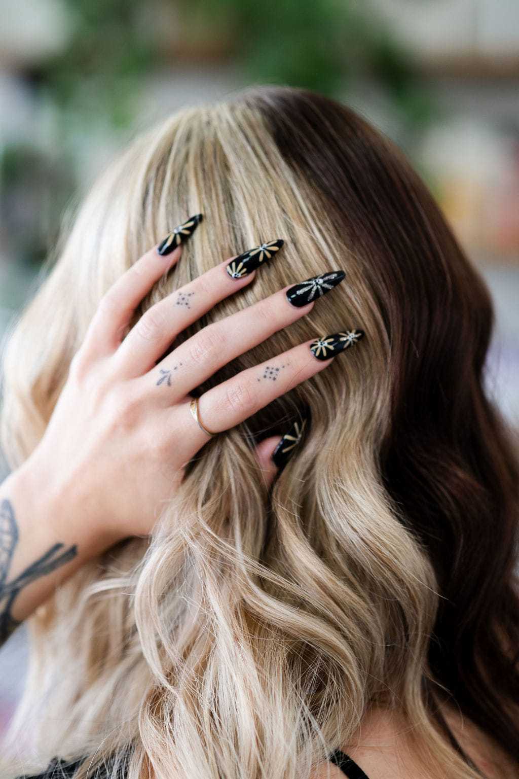 Woman with long, wavy blonde hair holding her head, displaying black nails with botanical designs.