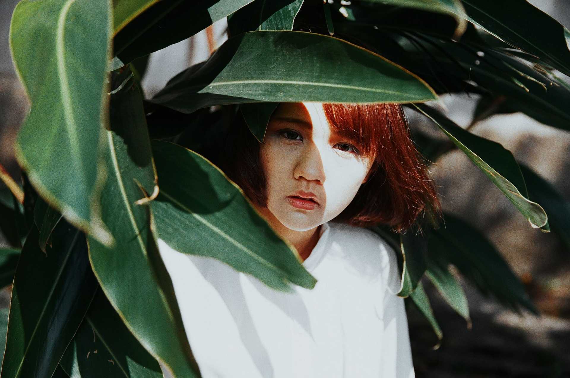 Woman partially hidden behind large green leaves, looking ahead with a neutral expression.