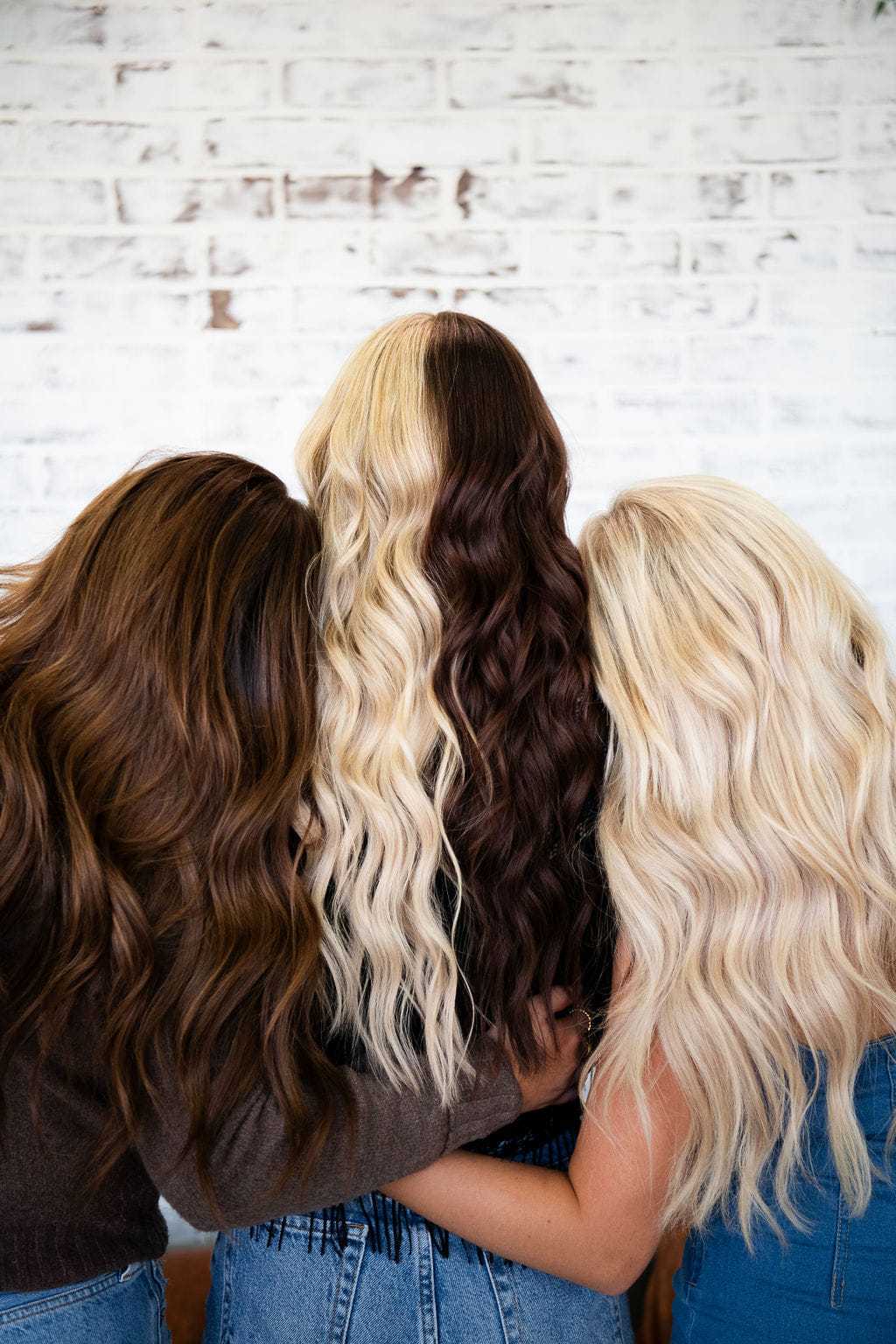 Three women with long, wavy hair in different shades, standing together against a white brick wall.